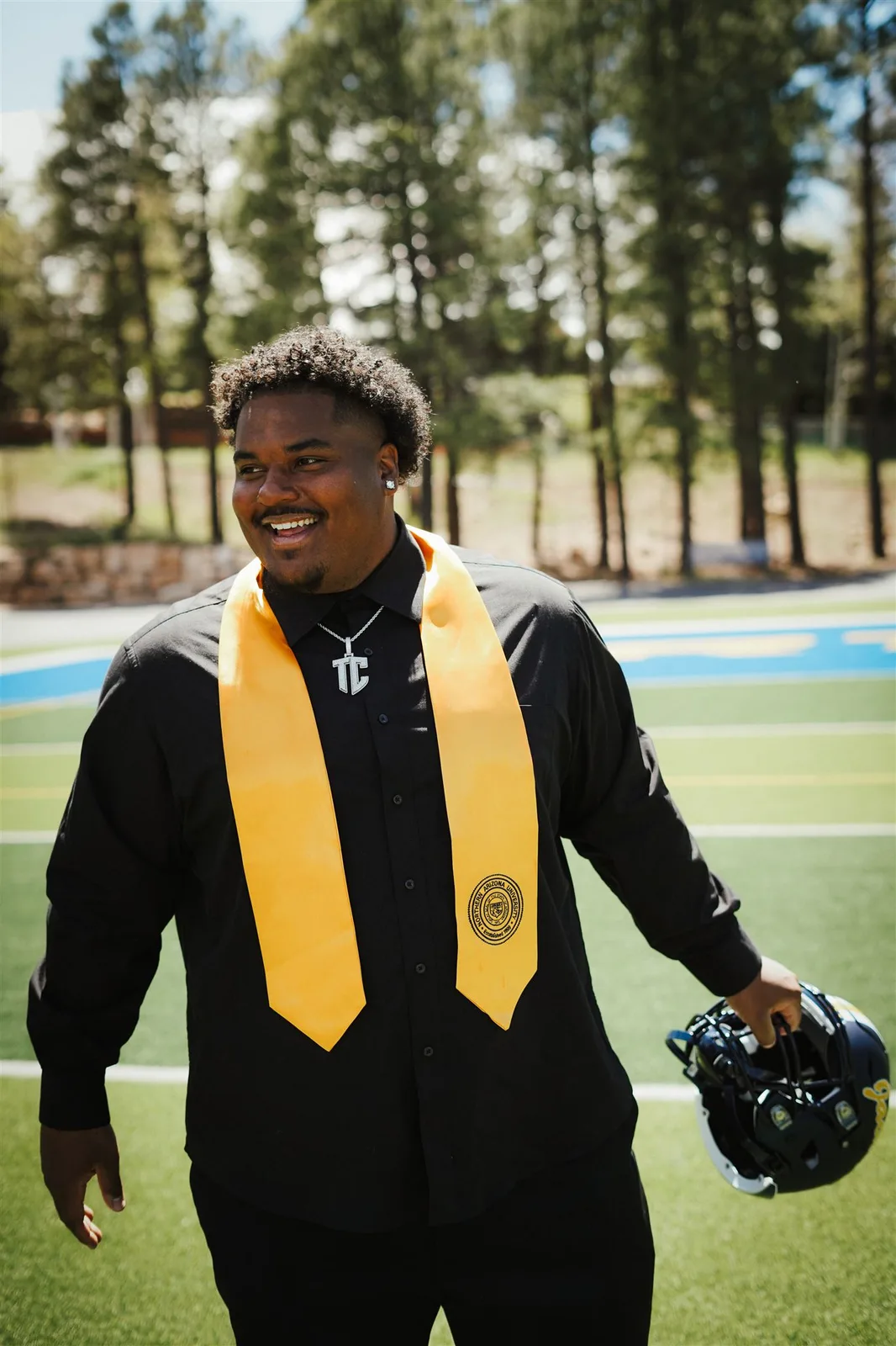 Terrance Caldwell in a black graduation gown and gold stole, holding a football helmet on a field lined with pine trees