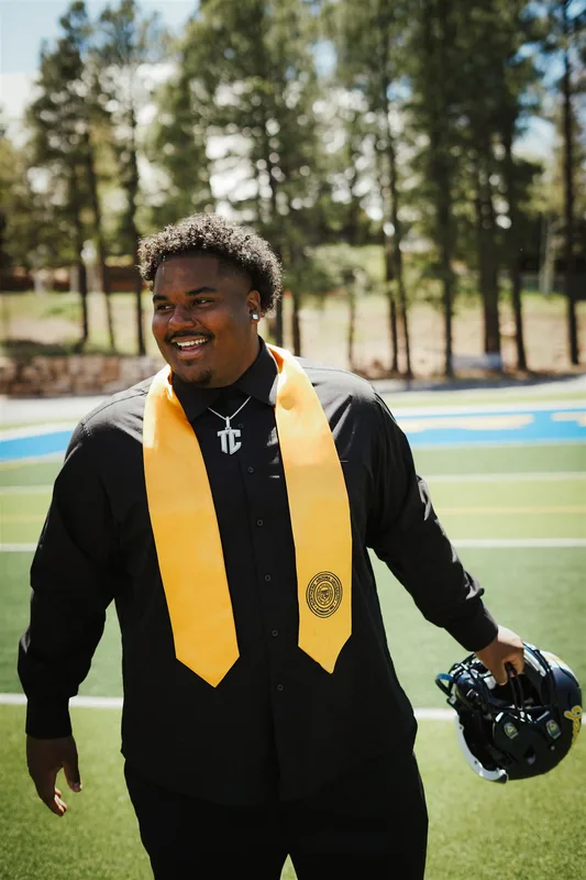 Terrance Caldwell in a black graduation gown and gold stole, holding a football helmet on a field lined with pine trees
